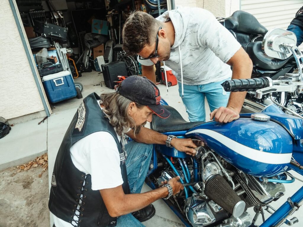 Two men focused on repairing a blue motorcycle inside a cluttered garage setting.