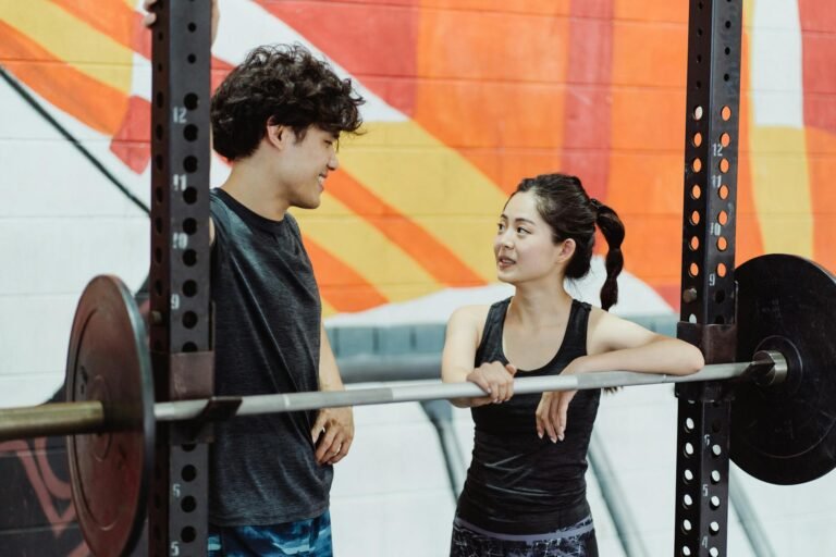 A young couple interacts at a gym while leaning on a barbell, promoting fitness and teamwork.