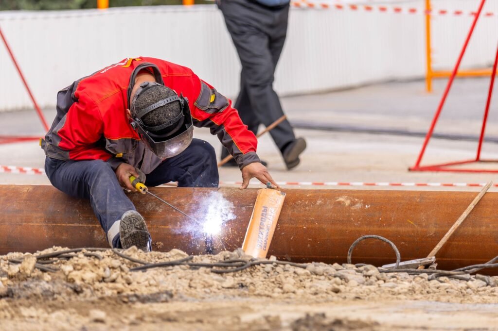 A skilled worker in safety gear welds a metal pipe at a construction site outdoors.