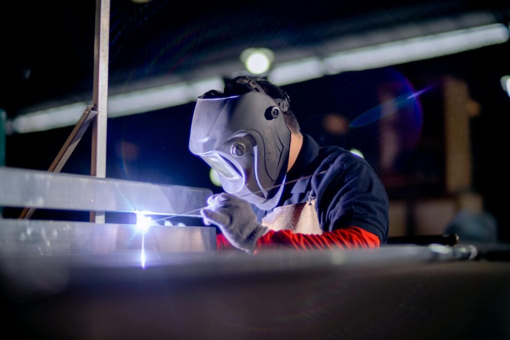 A welder in a protective mask works on metal in an industrial setting.