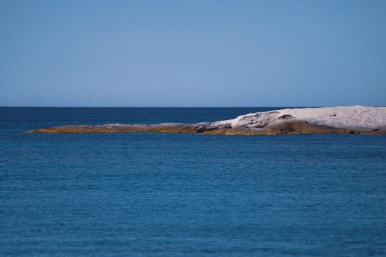 Serene view of a rocky island in a vast blue ocean under a clear sky, perfect for tranquil escapes.
