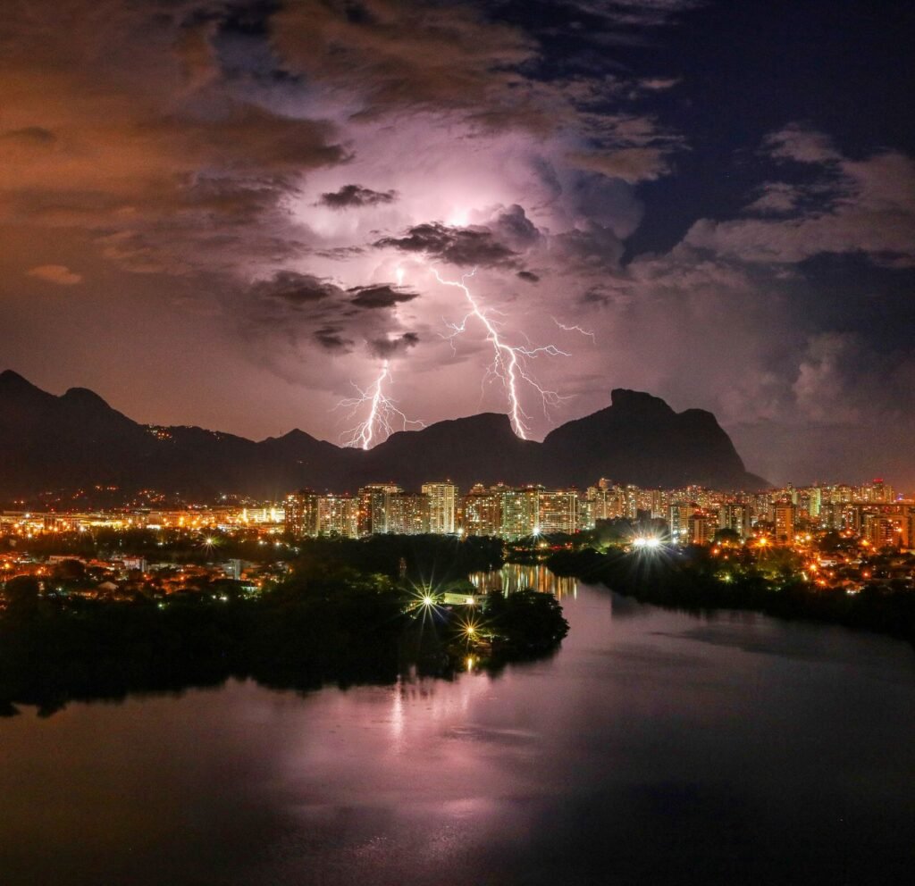 Picturesque view of bright lightning in gloomy sky above city buildings near river and mountains during thunderstorm at night
