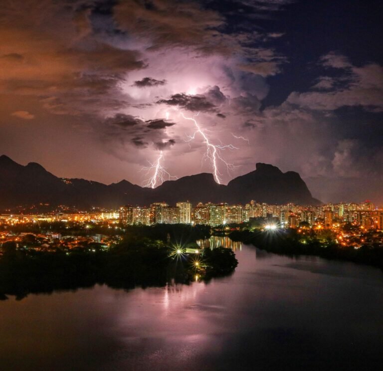 Picturesque view of bright lightning in gloomy sky above city buildings near river and mountains during thunderstorm at night